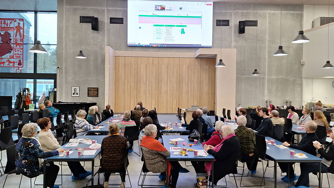 Yleisöä istuu pöytien äärellä ja katseet on suunnattu isolle, melko korkealla olevalle näytölle.