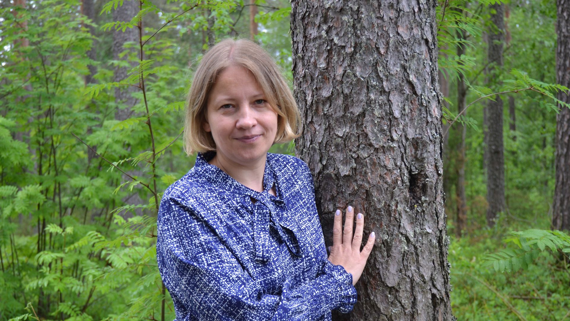 Siniseen pukeutunut naishenkilö nojaa mäntyyn, toinen käsi koskettaa runkoa.