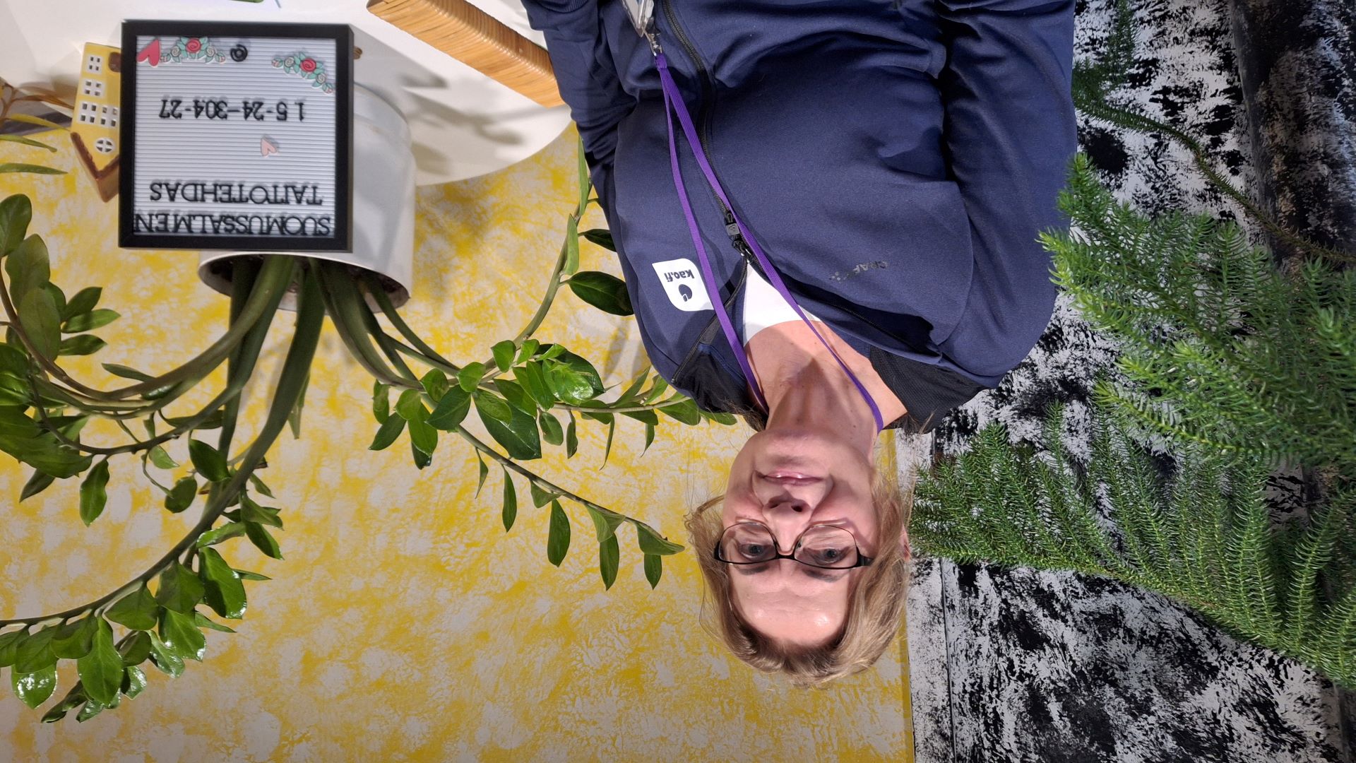 Silämälaispäinen henkilö puolilähikuvassa istuu tuolilla, takana näkyy kaksi viherkasvia. Oikealla olevalla pöydällä on Taitotehtaan kyltti. 