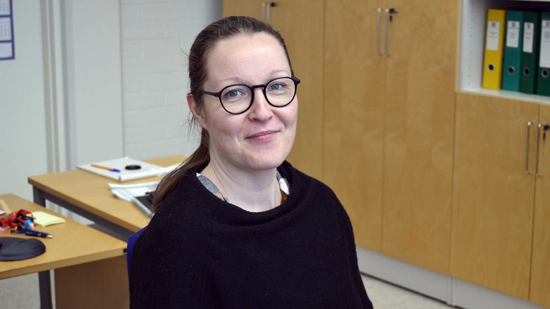 Lähikuvassa istuva ja hymyilevä henkilö, jolla yllään musta pusero ja tummasankaiset silmälasit. 