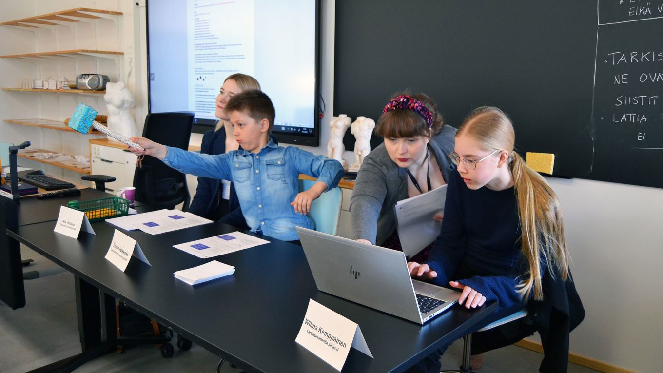 Lapsiparlamentin nuori puheenjohtaja osoittaa nuijalla saliin päin, hänen vierellään vaaleatukkainen nainen hymyilee, etualalla nuori kokouksen sihteeri istuu läppärin edessä ja hänen viereensä on kumartunut tummahiuksinen nainen papereita kädessään.