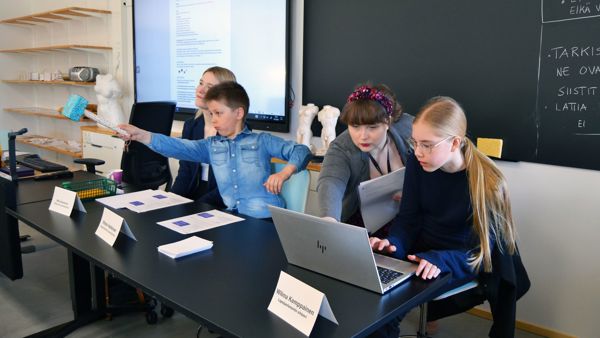 Lapsiparlamentin nuori puheenjohtaja osoittaa nuijalla saliin päin, hänen vierellään vaaleatukkainen nainen hymyilee, etualalla nuori kokouksen sihteeri istuu läppärin edessä ja hänen viereensä on kumartunut tummahiuksinen nainen papereita kädessään.