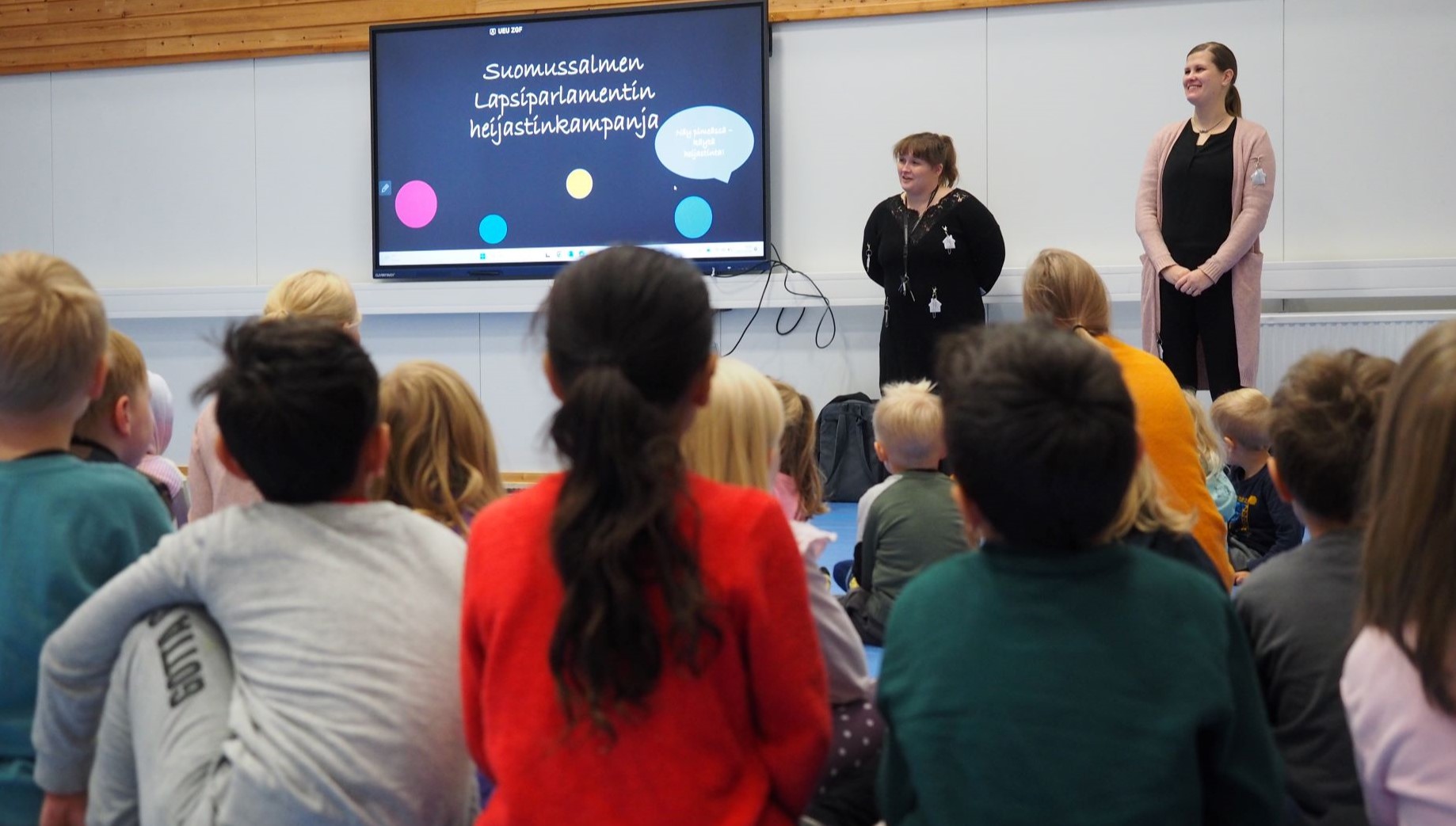 etualalla näkyy lapsia selin kameraan, taustalla kaksi aikuista seisoo ison näytön vieressä, näytön kuvassa lukee Suomussalmen lapsiparlamentin heijastinkampanja.