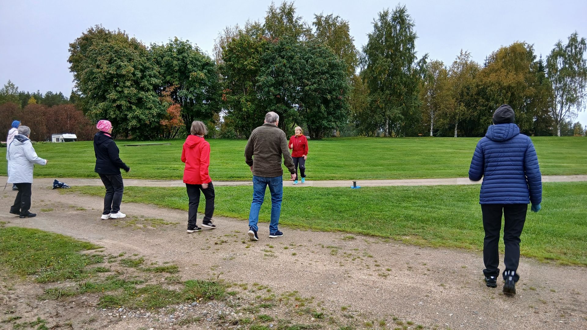 Kuusi henkilöä jumppaamassa puiston hiekkakäytävällä, takana näkyy ohjaaja.