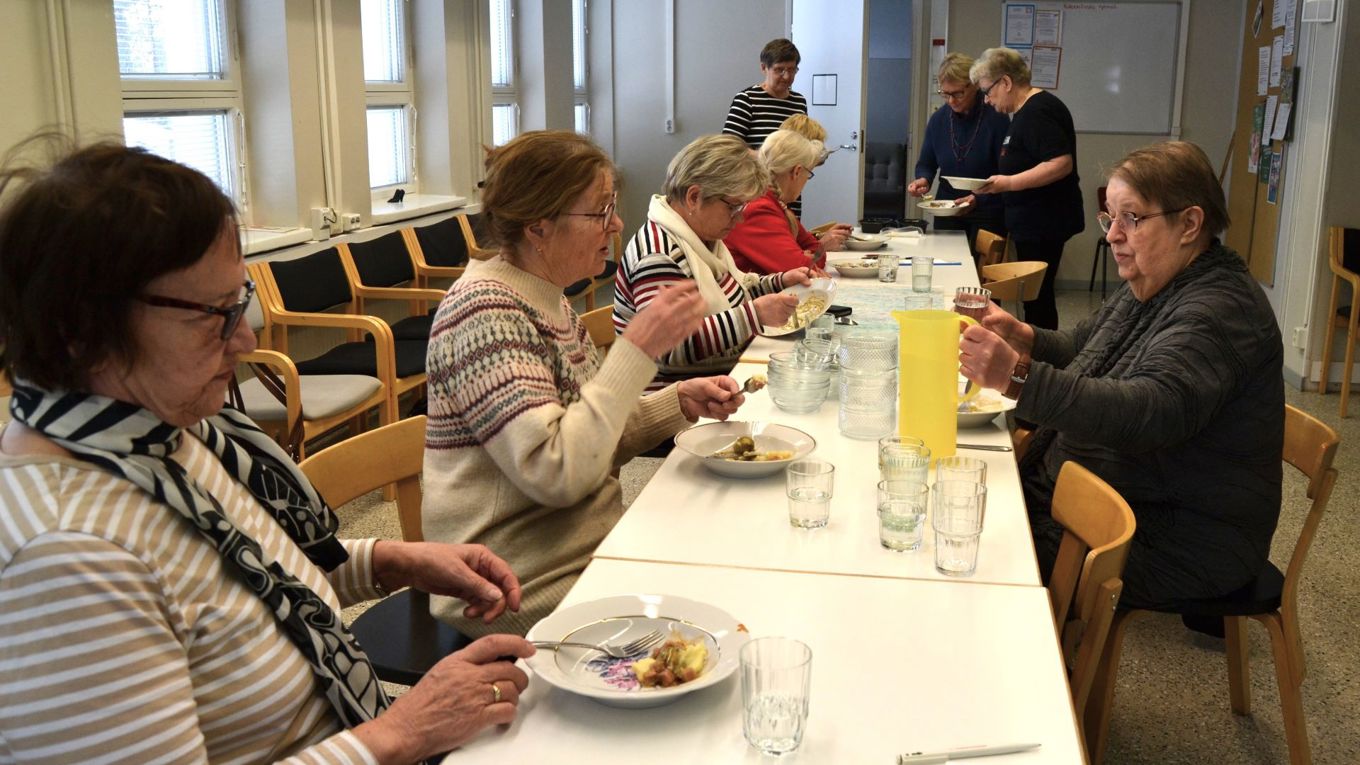 Kuusi henkilöä istuu  pitkän pöydän äärellä syömässä. Kolme henkilöä seisoo pöydän päässä ja ottaa ruokaa rasioista.