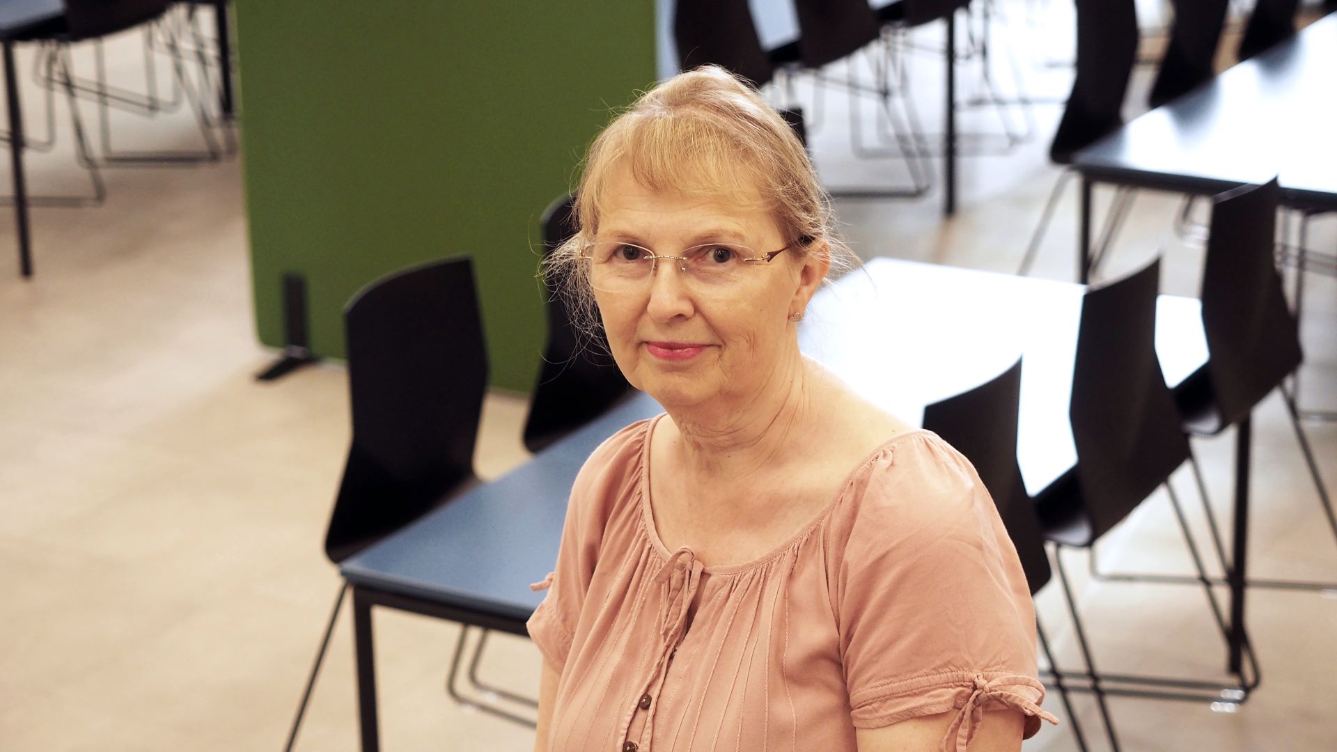 Puolilähikuvassa silmälasipäinen henkilö katsoo kuvaajaa. Taustalla näkyy koulun ruokalan pöytiä ja tuoleja. 