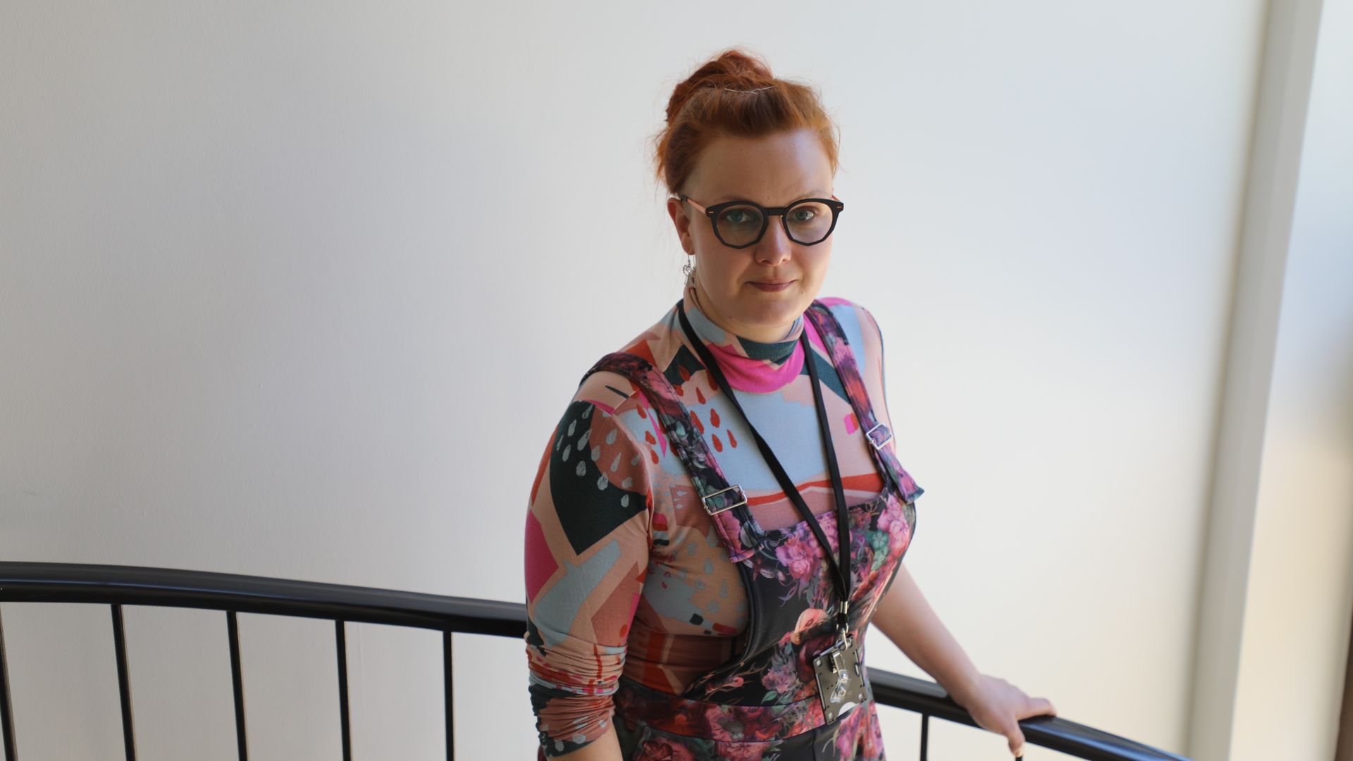 Kirjavaan paitaan pukeutunut nainen puolilähikuvassa seisoo portaikossa, vasen käsi pitelee kaidetta, seinä takana on vaalea.