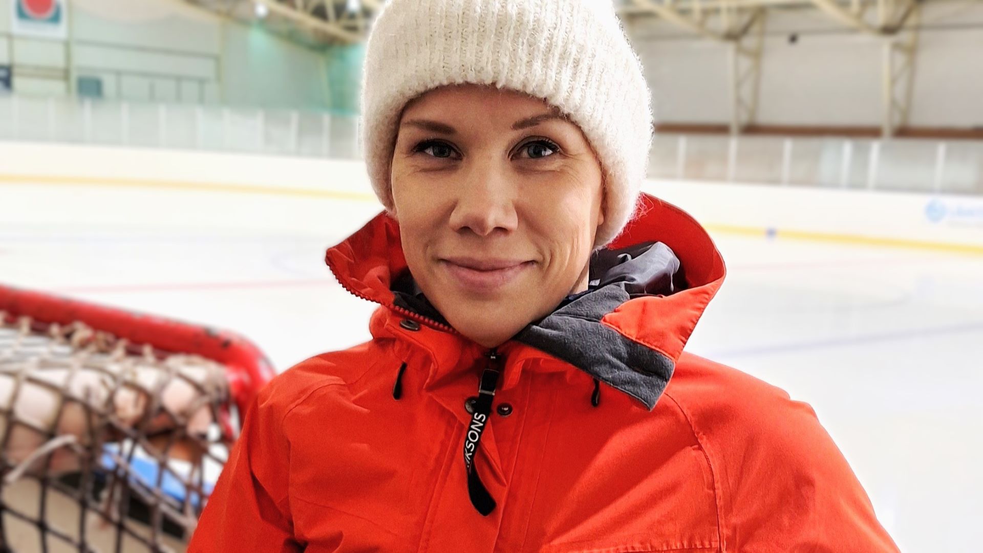 Valkoiseen pipoon ja oranssiin takkiin pukeutunut henkilö on lähikuvassa, taustanaan jääkiekkokaukalo, vasemmalla näkyy vähän jääkiekkomaalia. 