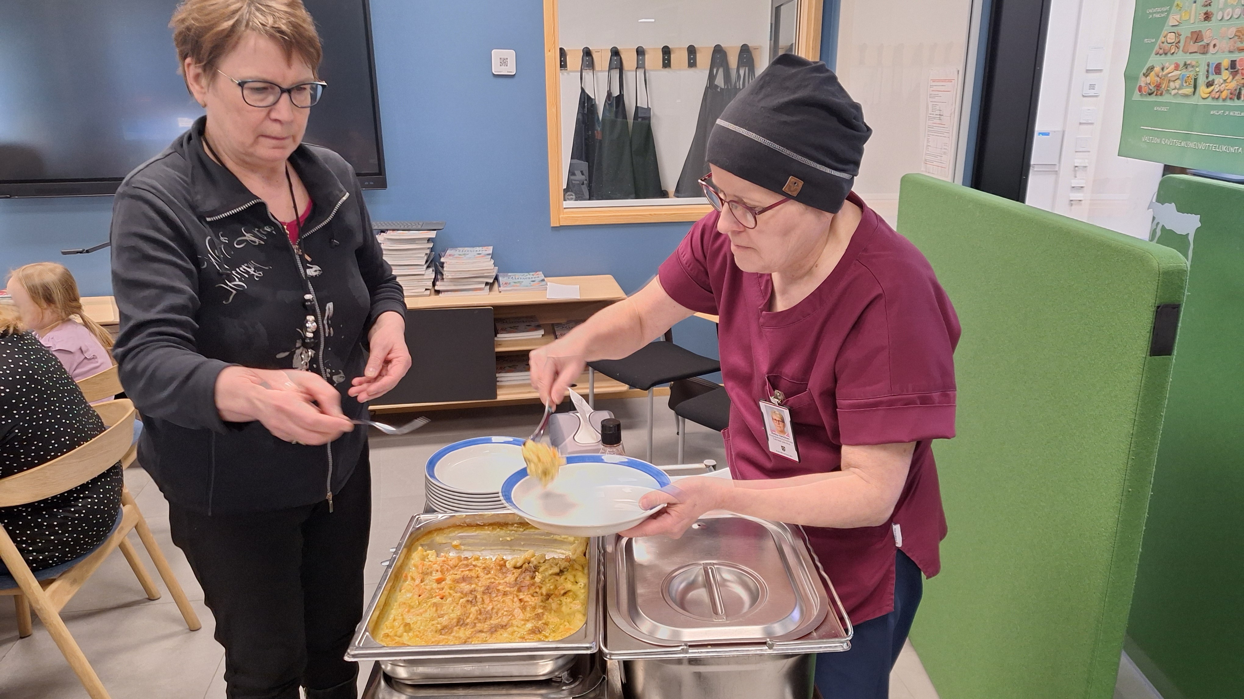 Kaksi henkilöä seisoo, toinen pitelee lautasta, jolle toinen laittaa ruokaa heidän välissää olevasta metallisesta astiasta.