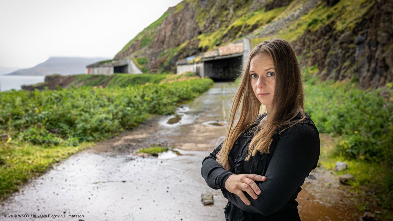 Kirjailija Satu Rämö islantilaisessa maisemassa. Rämö seisoo huonokuntoisella tiellä. Taustalla näkyy vuoren seinämää, jossa on tunnelin aukko. Taustalla näkyy myös vähän merta.