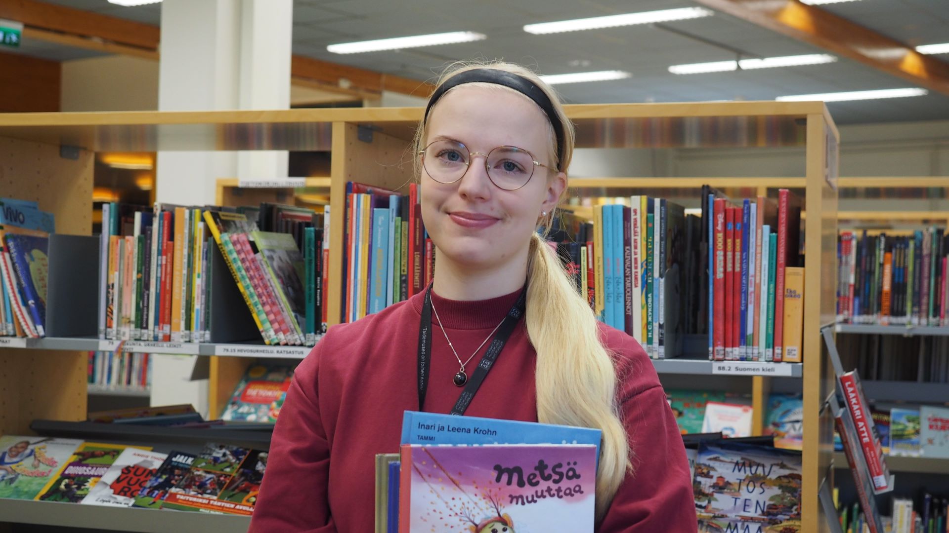 Hymyilevä vaaleatukkainen henkilö seisoo kirjaston kirjahyllyn edessä ja pitelee sylissään muutamia lastenkirjoja.