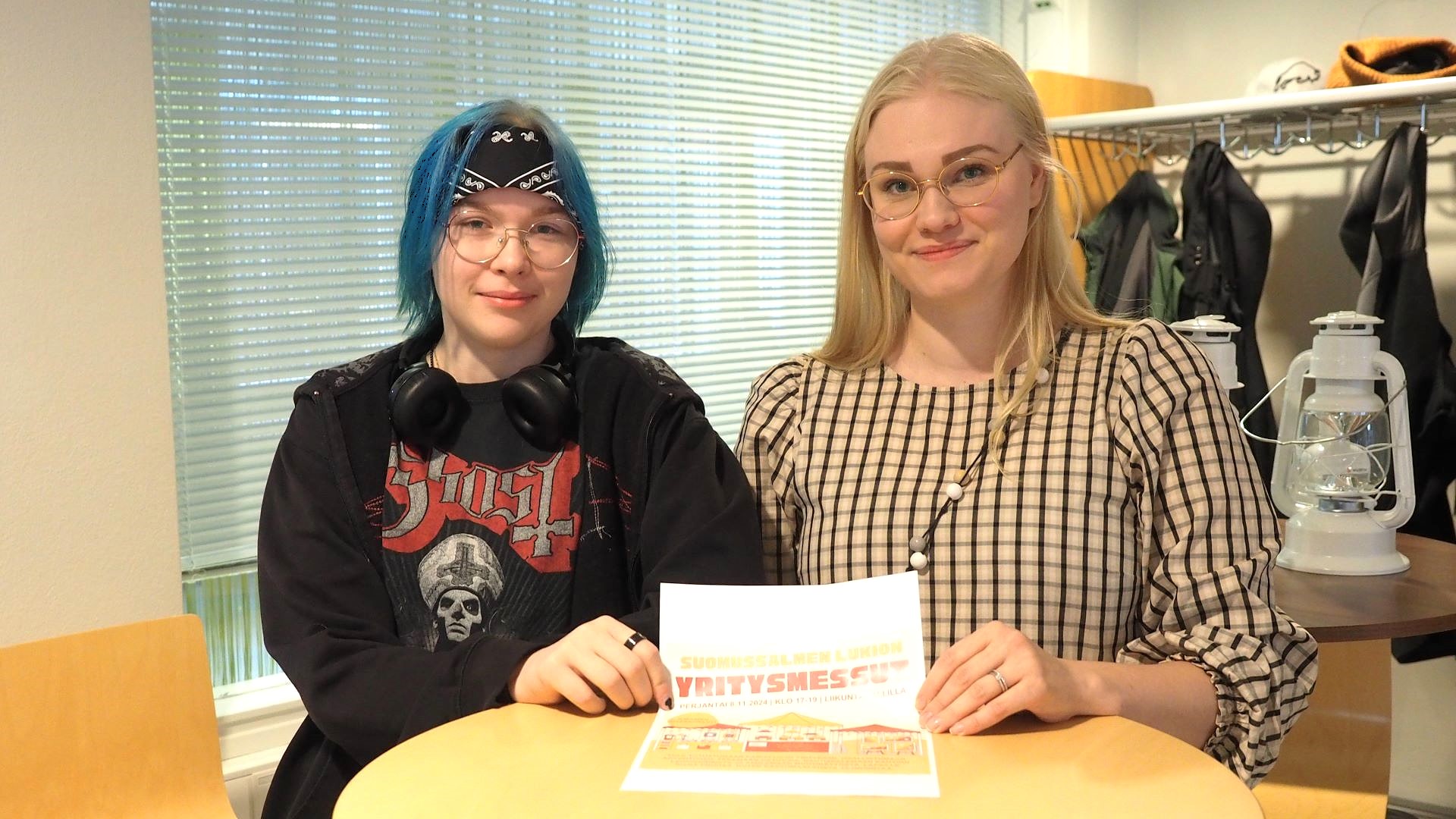 Kaksi henkilöä seisoo pienen pyöreän ja korkean pöydän takana, molemmat pitelevät samaa A4-kokoista mainosta yrittäjämessuista. 