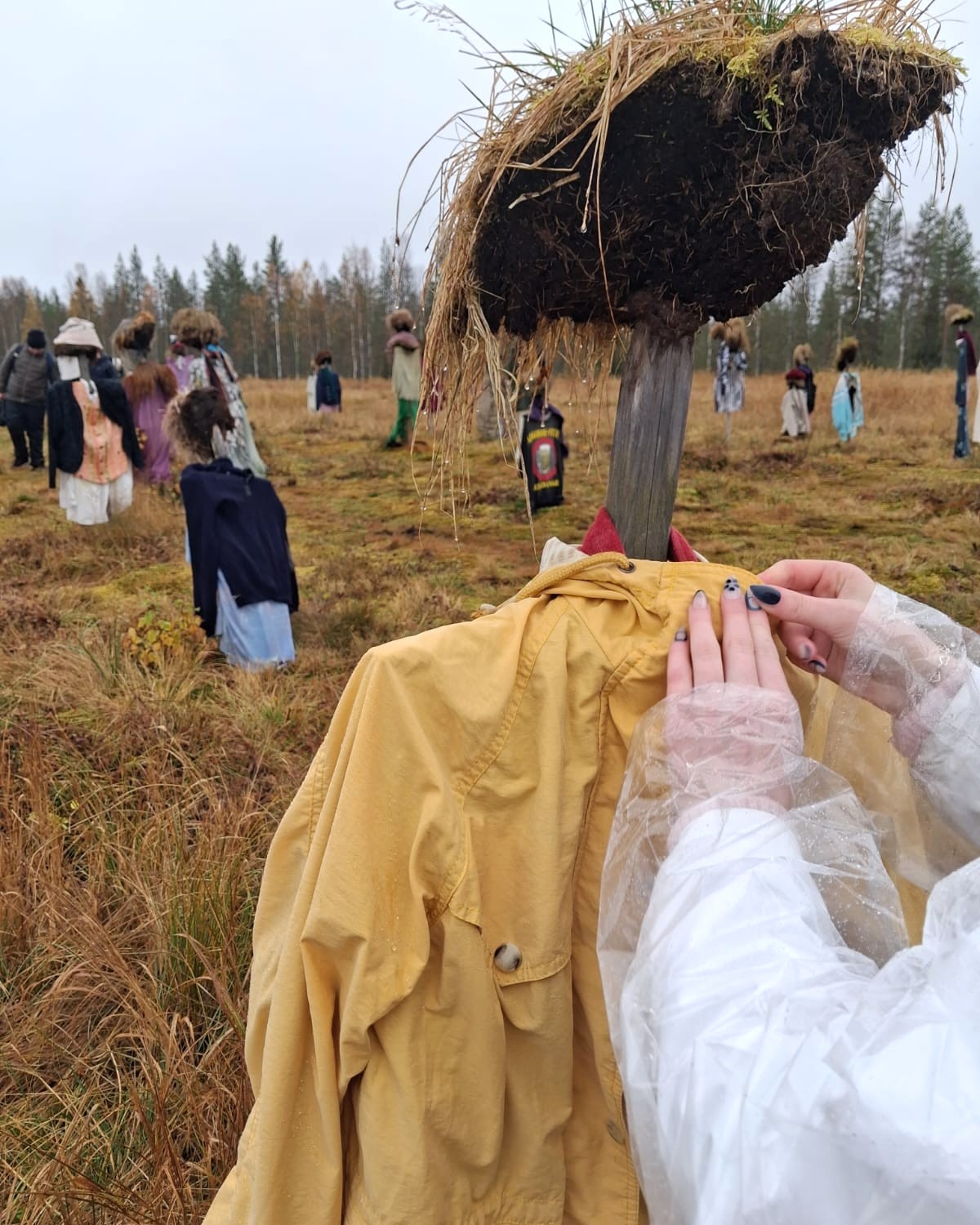Etuoikealla näkyvät valkoiseen takkiin ja läpinäkyvään sadetakkiin pukeutuneen henkilön kädet, jotka asettelevat keltaista anorakkia hahmon ylle. Taustalla näkyy joitakin hahmoja.