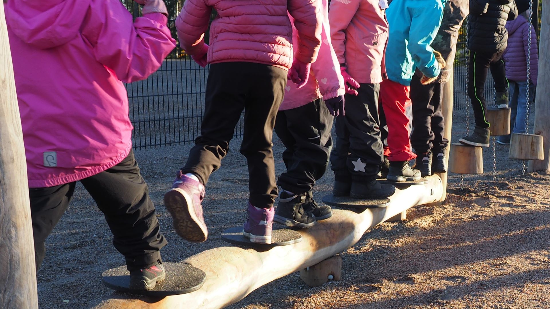 Kuvituskuvassa näkyy lasten jalkoja ja värikkäitä takkeja, lapsia seisoo tai kulkee paksun lankun päällä.