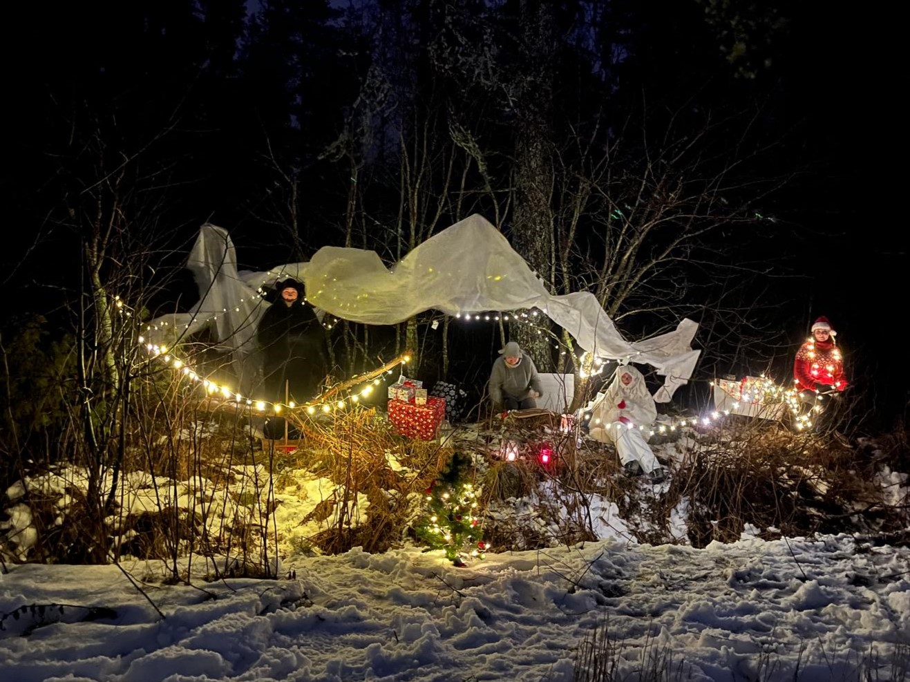 Ulkona lumisessa maastossa, takana metsää, on valkoinen telttamainen katos, muutama henkilö näkyy, kauniita kirkkaita jouluvaloja ja lahjapaketteja kauniissa asetelmassa.