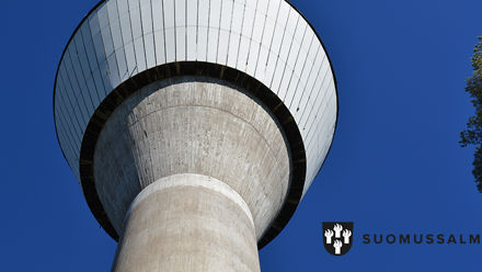 Vesitornin yläosa näkyy kirkkaansinistä taivasta vasten. Oikealla männyn latvaa.