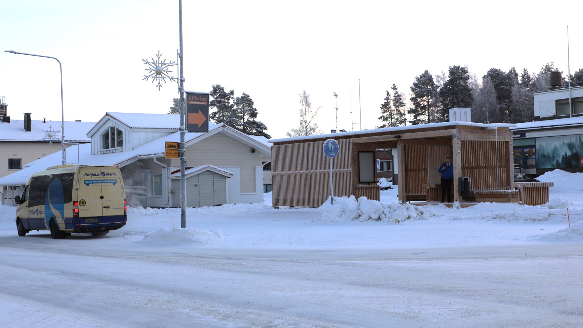 Talvella otettu kuva risteyksen takaa, vasemmalla pieni linja-auto ja jalkakäytävän toisella puolella on pystyistä lehtikuusirimoista tehty katos.