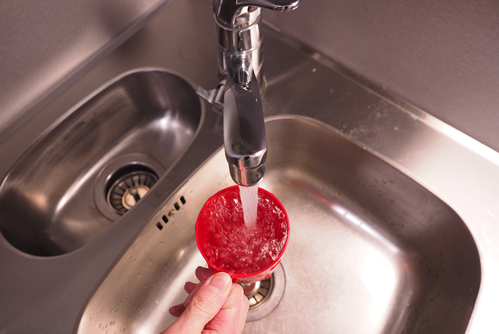 Kuvituskuvassa käsi pitelee punaista muovimukia valuvan vesihanan alla.