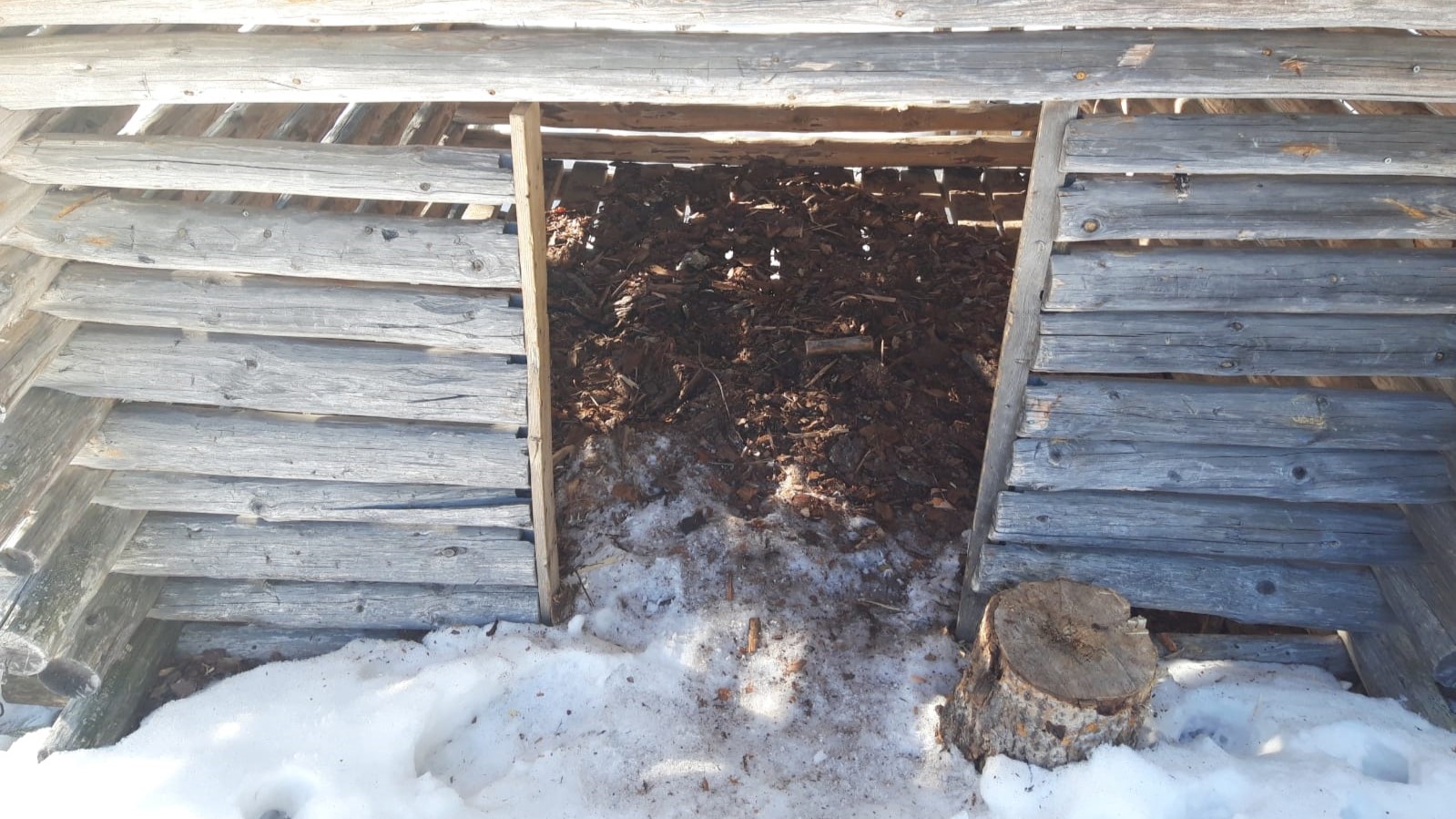 hirsirakenteisen puukatoksen oviaukossa näkyy tyhjennetty liiteri, edustalla on lunta ja oviaukon vieressä hakkuupölkky.