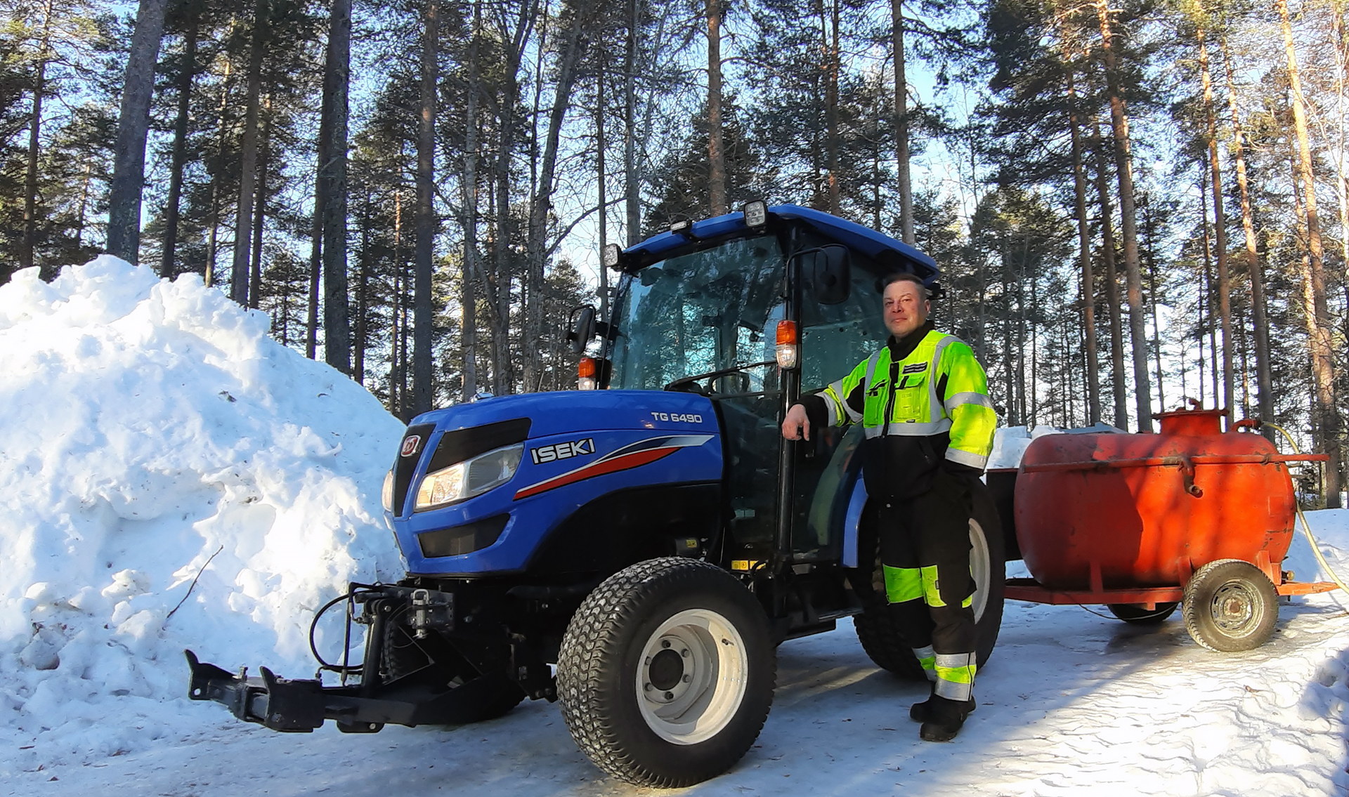 Työasuun pukeutunut henkilö poseeraa sinisen traktorin vierellä, traktorin perässä on punainen vesisäiliö pyörien päällä. Vasemmalla on iso lumikasa, taustalla mäntymetsää.
