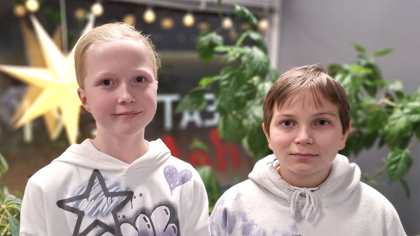 Kaksi kymmenvuotiasta lasta vierekkäin puolilähikuvassa. Molemmilla on yllään vaalea huppari. Taustalla näkyy iso ikkuna ja valaistu joulutähti, lisäksi viherkasveja.
