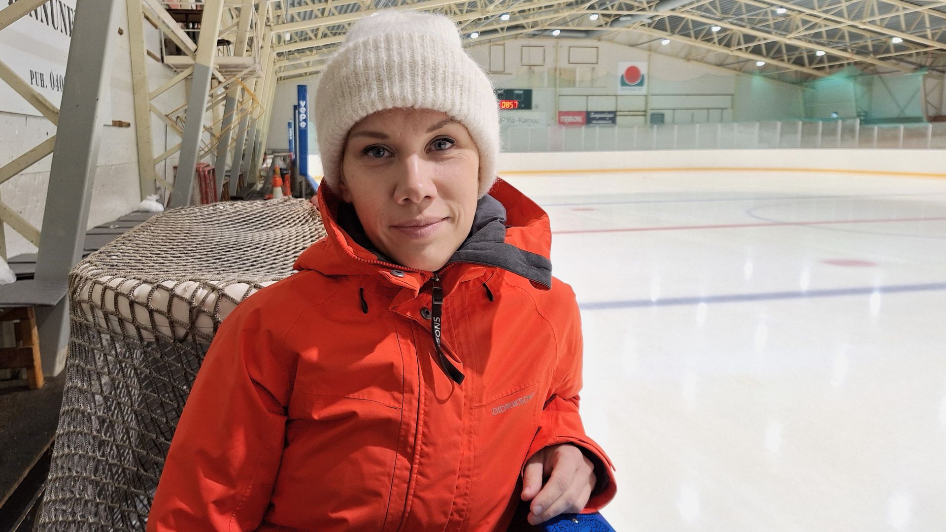 Punaiseen takkiin ja valkoiseen pipoon pukeutunut henkilö puolilähikuvassa nojaa kyynärpäällä jääkiekkokaukalon laitaan, kaukalon ulkpuolella. 