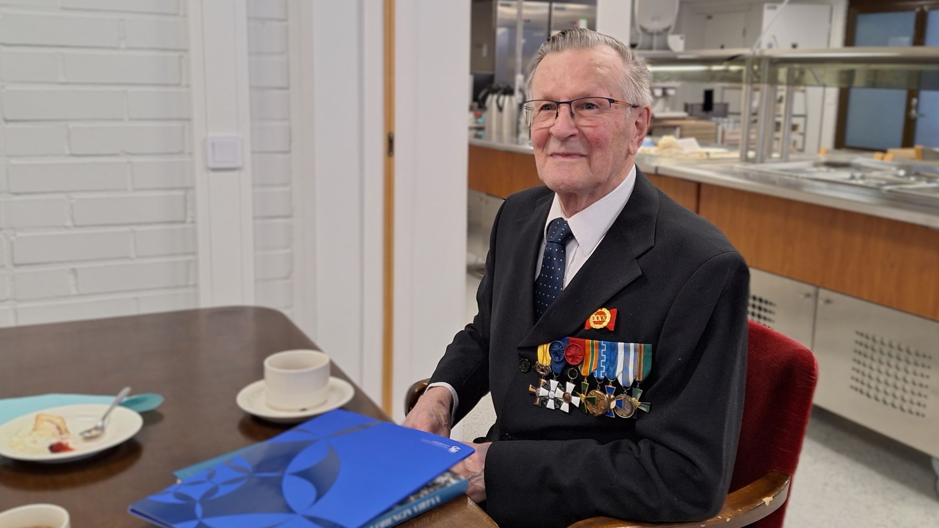 Varttunut mies tummassa puvussa, jonka rintapielessä on useita kunniamerkkejä, istuu pöydän äärellä edessään sininen kansio ja kahvikuppi.