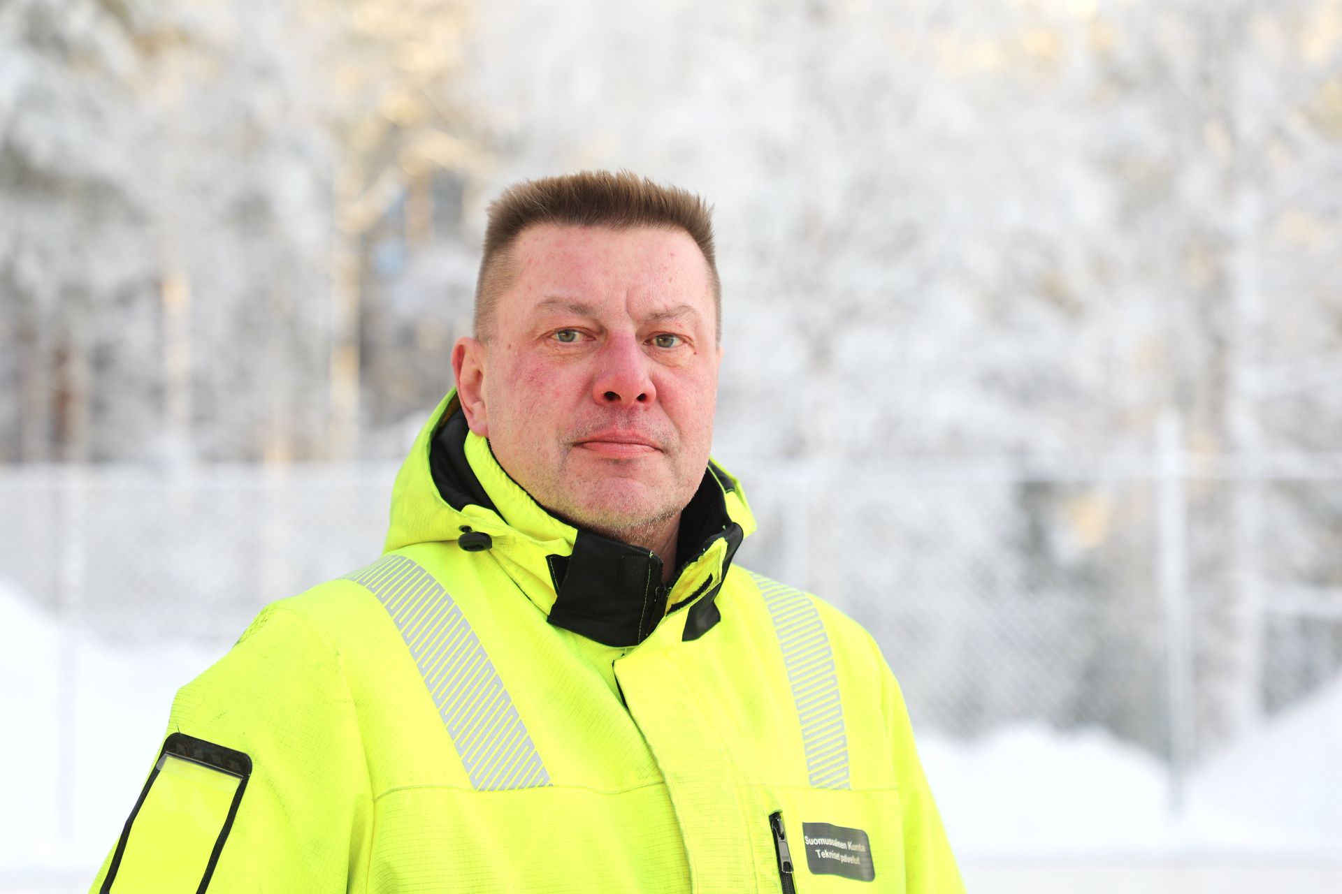 Keltaiseen työtakkiin pukeutunut mies lähikuvassa, taustalla lumista metsää ja huurteessa olevaa metalliaitaa. 