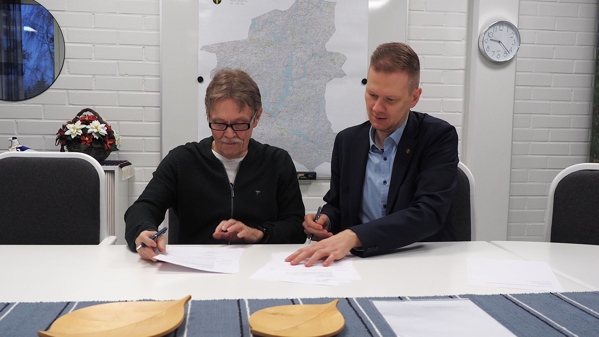 Kaksi henkilöä istuu pöydän äärellä, molemmat hypistelevät papereita ja molemmilla on kynä kädessään.