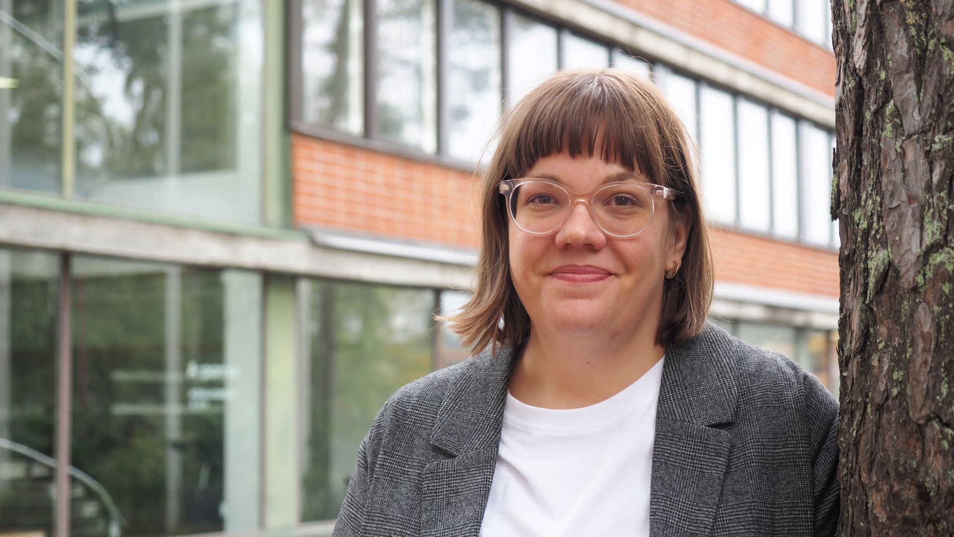 Lähikuvassa hymyilevä silmälasipäinen nainen lähikuvassa seisoo ison männyn vieressä, puusta näkyy vain vähän runkoa. Taustalla näkyy kunnantalon ikkunoita ja punatiilistä seintää.