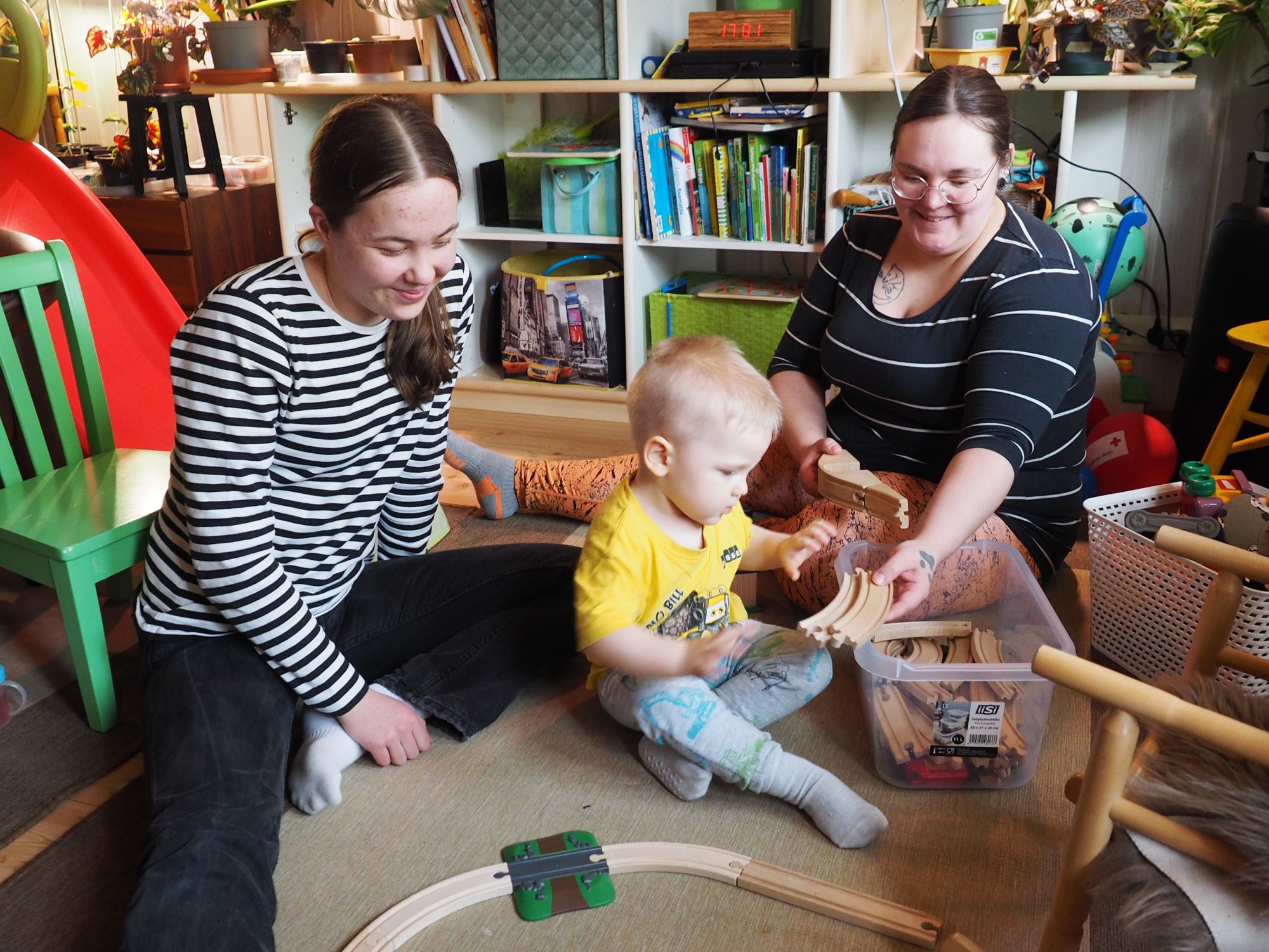 Olohuoneen lattialla istuu kolme henkilöä, joista yksi on lapsi, joka leikkii lelujunalla.