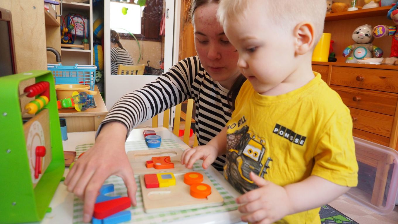 Lastenhuoneessa keltaisessa paidassa vaaleatukkainen pikkupoika leikkii värikkäällä palapelillä, hänen takanaan on raidalliseen paitaan pukeutunut nuori nainen, jonka eteen ojennettu käsi tarttuu punaiseen palaan.