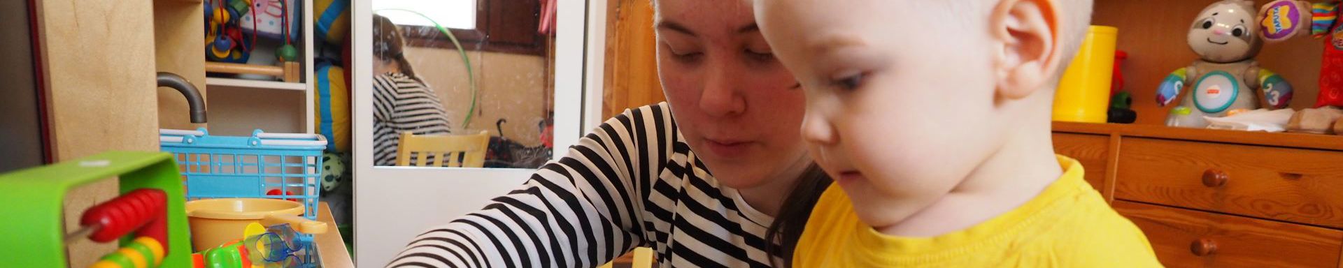 Nuori nainen ja keltaiseen paitaan puettu pikkulapsi pöydän äärellä leikkivät värikkäillä muoviesineillä.