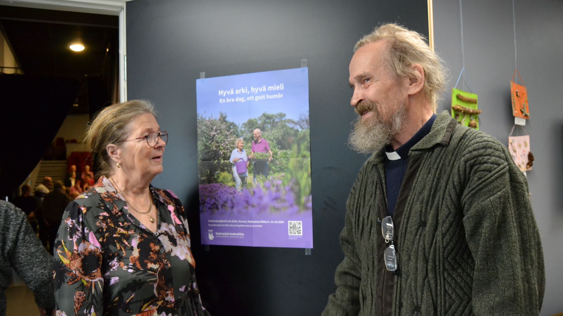 Kaksi henkilöä seisoo tummanharmaan oven vieressä, heidän välissään oveen on kiinnitetty Vanhustenviikon juliste. 